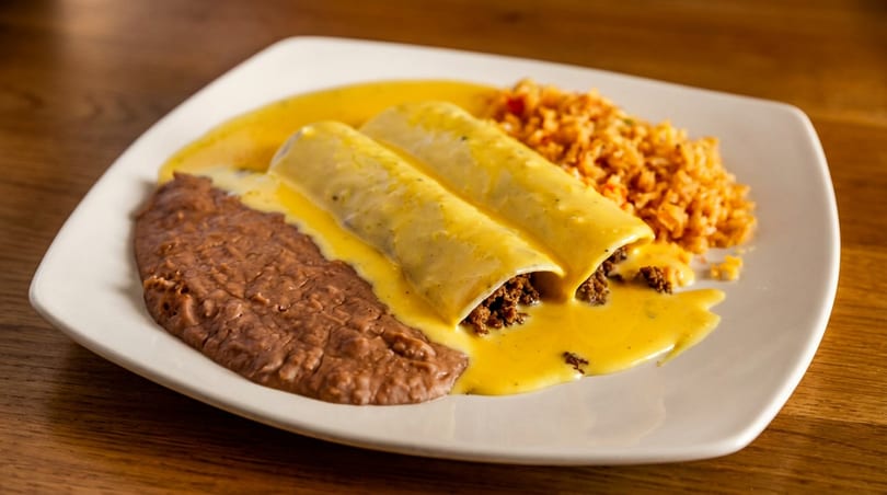 Two beef enchiladas covered in yellow cheese sauce, with refried beans and rice on a white plate.