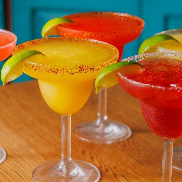 Five colorful margaritas, some red and some yellow, garnished with lime wedges and salt on a wooden table.
