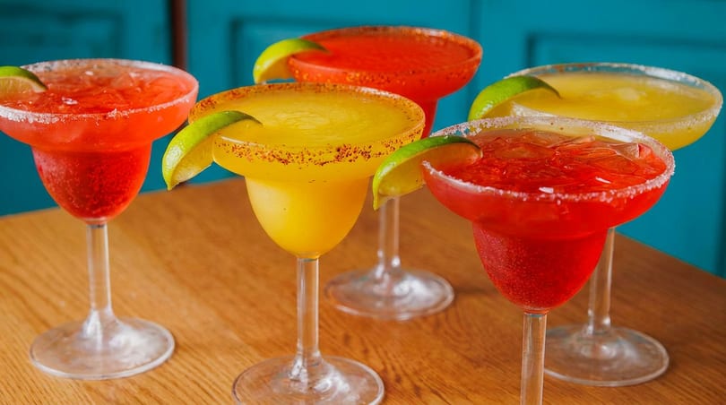 Five colorful margaritas, some red and some yellow, garnished with lime wedges and salt on a wooden table.