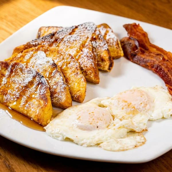 A white plate with French toast, powdered sugar, syrup, two fried eggs, and crispy bacon.