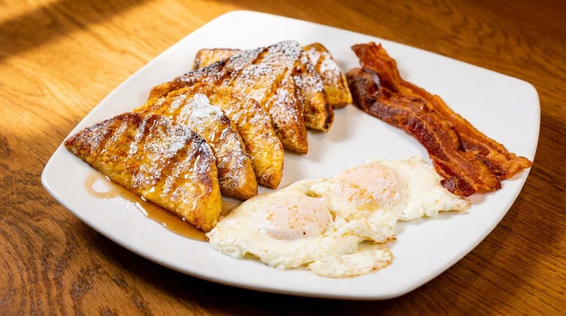 A white plate with French toast, powdered sugar, syrup, two fried eggs, and crispy bacon.