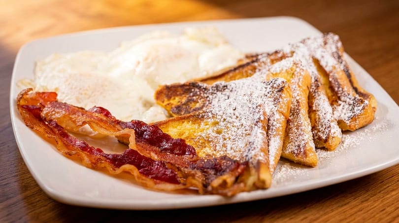 French toast with powdered sugar, crispy bacon, and scrambled eggs on a white plate.