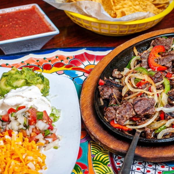Sizzling beef fajitas with onions and peppers, alongside beans, guacamole, salsa, and chips.
