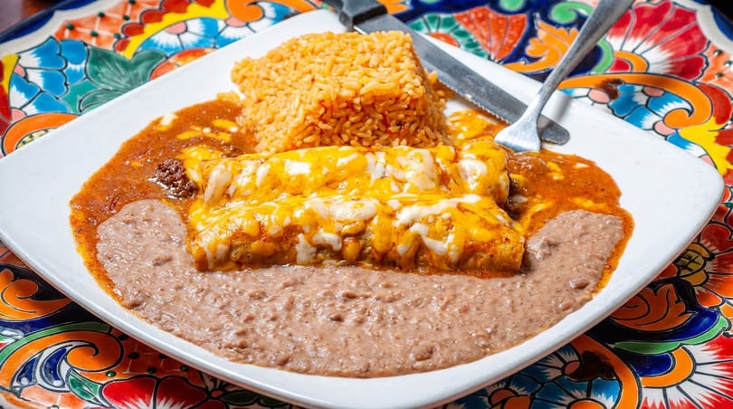 Cheesy enchiladas with red sauce, refried beans, and rice on a white plate with a colorful placemat.