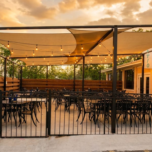 Outdoor restaurant patio with string lights, shade sails, and empty tables under a sunset sky.