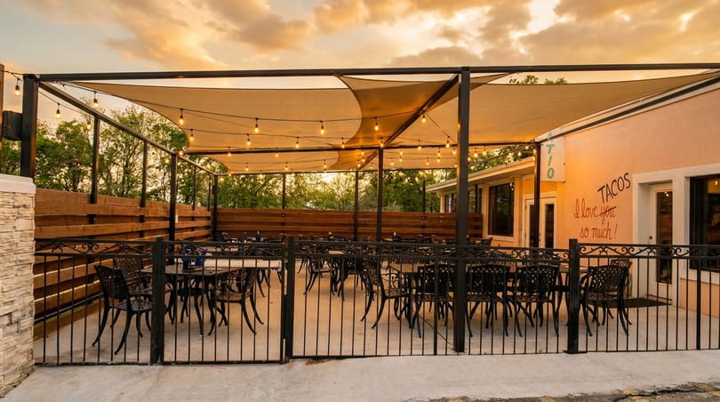 Outdoor restaurant patio with string lights, shade sails, and empty tables under a sunset sky.
