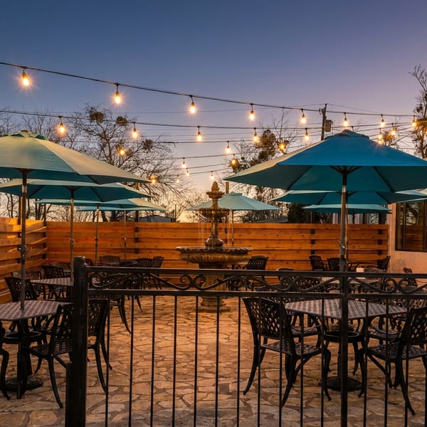 An empty restaurant patio at dusk with string lights, teal umbrellas, and a stone fountain.