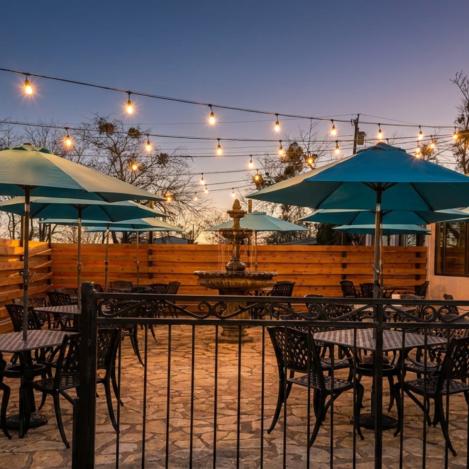 An empty restaurant patio at dusk with string lights, teal umbrellas, and a stone fountain.