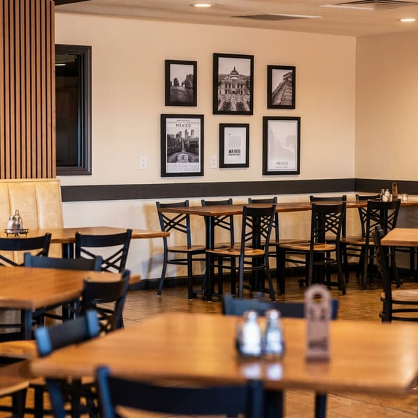 A restaurant dining room with wooden tables, black chairs, a long booth, and framed Mexico art.