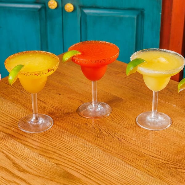 Five colorful margaritas, some red and some yellow, with lime wedges on a wooden table.