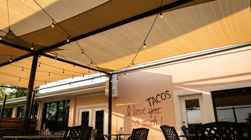 Outdoor patio with string lights under a beige canopy, and a wall with "TACOS I love you so much!" written on it.