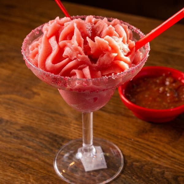 A vibrant red frozen margarita with two straws and a side of salsa on a wooden table.