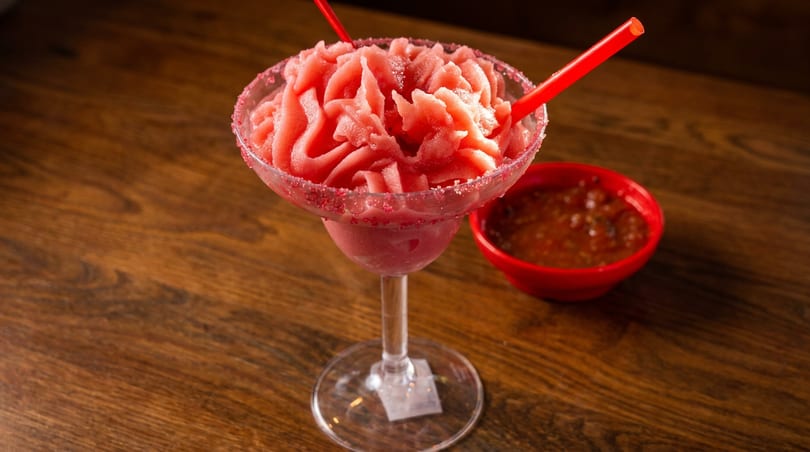 A vibrant red frozen margarita with two straws and a side of salsa on a wooden table.
