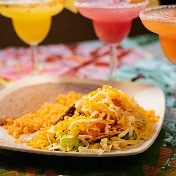 A plate of tacos, rice, and beans with three colorful margaritas in the background.