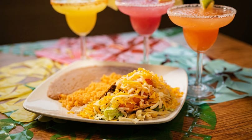 A plate of tacos, rice, and beans with three colorful margaritas in the background.
