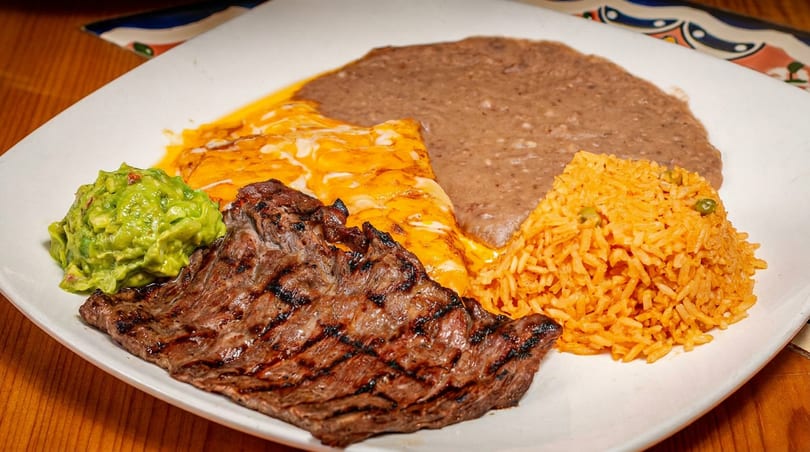 Grilled steak, guacamole, enchiladas, refried beans, and rice on a white plate.
