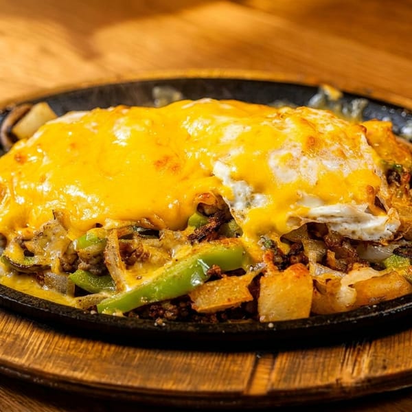 Sizzling skillet of breakfast potatoes, peppers, onions, and meat, topped with melted cheese and a fried egg.