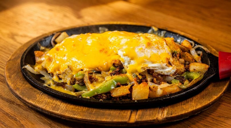 Sizzling skillet of breakfast potatoes, peppers, onions, and meat, topped with melted cheese and a fried egg.